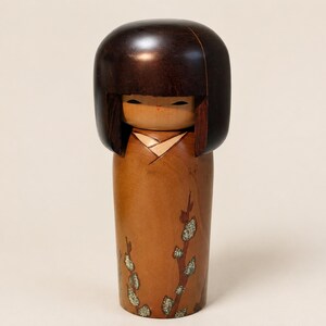 K&ouml;nnte beinhalten: Eine h&ouml;lzerne Kokeshi-Puppe mit einem glatten, dunkelbraunen, abgerundeten Kopf und einem hellbraunen zylindrischen K&ouml;rper. Der K&ouml;rper ist mit einem gemalten Zweigmotiv verziert, das kleine, texturierte gr&uuml;ne und wei&szlig;e Knospen zeigt.