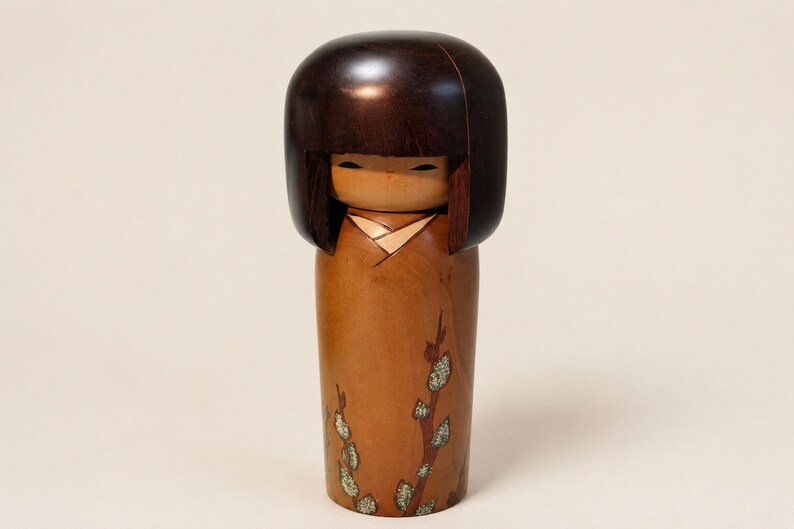 K&ouml;nnte beinhalten: Eine h&ouml;lzerne Kokeshi-Puppe mit einem glatten, dunkelbraunen, abgerundeten Kopf und einem hellbraunen zylindrischen K&ouml;rper. Der K&ouml;rper ist mit einem gemalten Zweigmotiv verziert, das kleine, texturierte gr&uuml;ne und wei&szlig;e Knospen zeigt.
