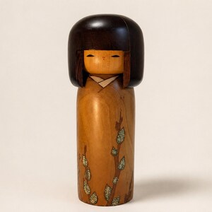 K&ouml;nnte beinhalten: Eine h&ouml;lzerne Kokeshi-Puppe mit einem glatten, dunkelbraunen, abgerundeten Kopf mit Bob-Frisur und einem hellbraunen zylindrischen K&ouml;rper. Der K&ouml;rper ist mit einem gemalten Muster aus d&uuml;nnen braunen Zweigen und kleinen, hellgr&uuml;nen, texturierten Blattgruppen verziert.