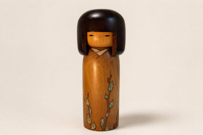 K&ouml;nnte beinhalten: Eine h&ouml;lzerne Kokeshi-Puppe mit einem glatten, dunkelbraunen, abgerundeten Kopf mit Bob-Frisur und einem hellbraunen zylindrischen K&ouml;rper. Der K&ouml;rper ist mit einem gemalten Muster aus d&uuml;nnen braunen Zweigen und kleinen, hellgr&uuml;nen, texturierten Blattgruppen verziert.