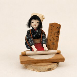 May include: A Japanese doll with black hair and a beige head covering, wearing a dark patterned kimono with a red apron. The doll is positioned behind a wooden board with a rolling pin and a circular dough piece, next to a wooden sign with black text.