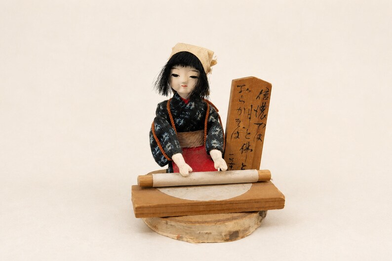 May include: A Japanese doll with black hair and a beige head covering, wearing a dark patterned kimono with a red apron. The doll is positioned behind a wooden board with a rolling pin and a circular dough piece, next to a wooden sign with black text.