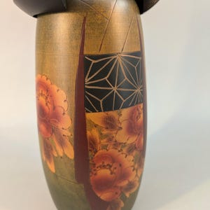 May include: A wooden Kokeshi doll featuring a cylindrical body with a muted olive green base color. The surface is decorated with orange and red floral patterns and a black rectangular panel containing a white geometric line design.