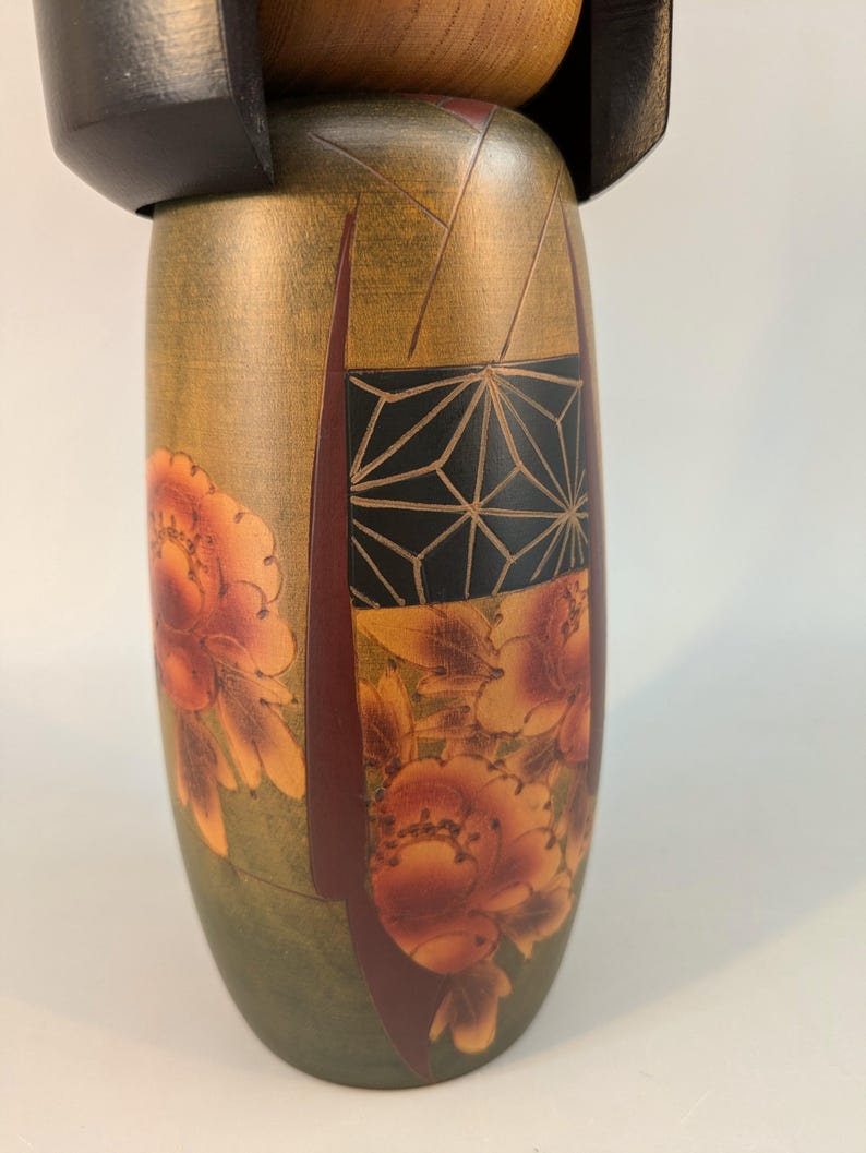 May include: A wooden Kokeshi doll featuring a cylindrical body with a muted olive green base color. The surface is decorated with orange and red floral patterns and a black rectangular panel containing a white geometric line design.