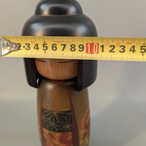 May include: A wooden Kokeshi doll with a smooth black head and a natural wood face featuring closed eyes. The body is painted in olive green with a black geometric patterned obi and red floral accents. A yellow measuring tape shows a width of 3.5 inches.