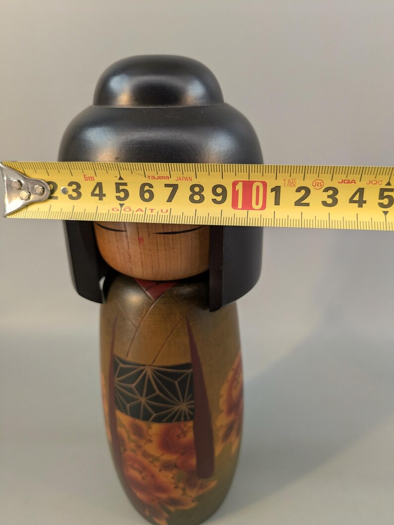May include: A wooden Kokeshi doll with a smooth black head and a natural wood face featuring closed eyes. The body is painted in olive green with a black geometric patterned obi and red floral accents. A yellow measuring tape shows a width of 3.5 inches.