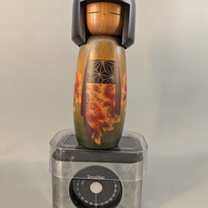 May include: A wooden Kokeshi doll with a black bob hairstyle and a natural wood face, featuring a body painted with green, orange floral patterns, and a black geometric design. The doll stands on a silver-colored Terraillon kitchen scale.