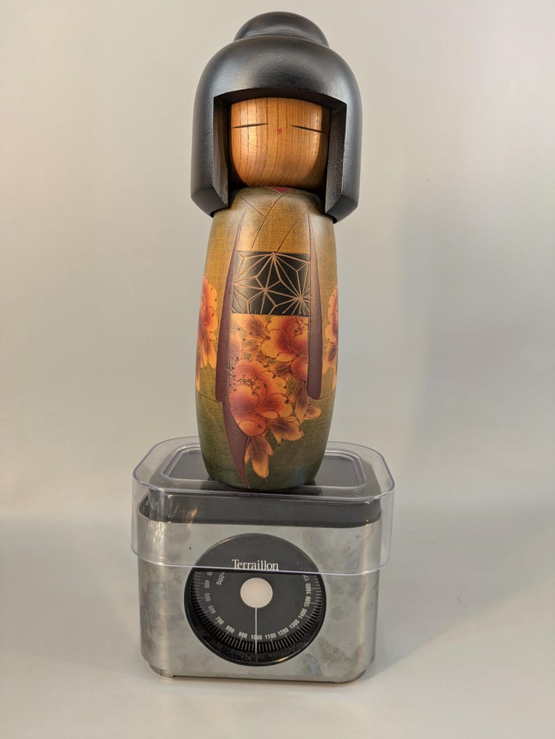 May include: A wooden Kokeshi doll with a black bob hairstyle and a natural wood face, featuring a body painted with green, orange floral patterns, and a black geometric design. The doll stands on a silver-colored Terraillon kitchen scale.