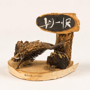 May include: A decorative wooden sculpture featuring a brown, textured wild boar figure positioned on a light-coloured wood base. A vertical wooden post supports an oval sign with a black centre and white Japanese characters painted on the surface.