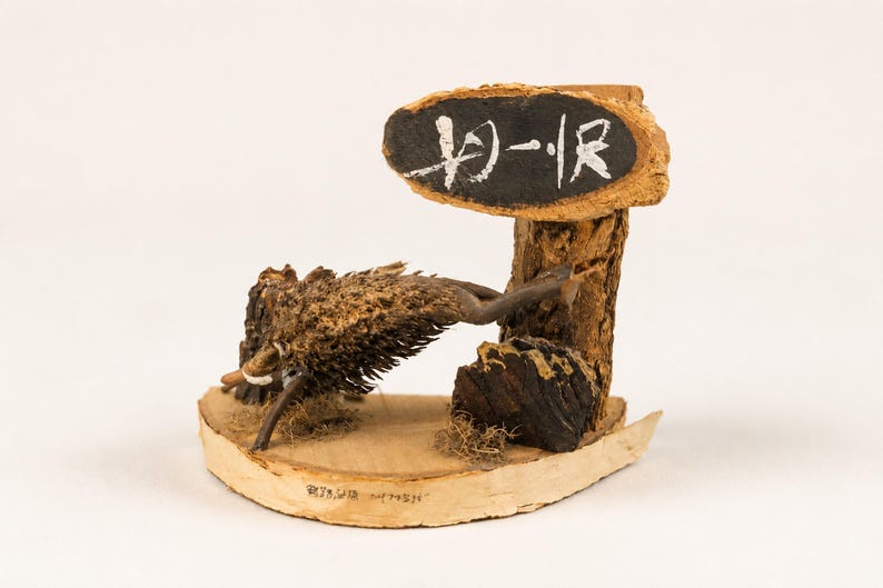 May include: A decorative wooden sculpture featuring a brown, textured wild boar figure positioned on a light-coloured wood base. A vertical wooden post supports an oval sign with a black centre and white Japanese characters painted on the surface.