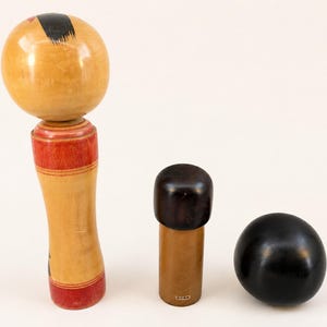 K&ouml;nnte beinhalten: Drei Holzobjekte, darunter eine gro&szlig;e Kokeshi-Puppe mit rundem Kopf und rot bemaltem K&ouml;rper, ein kleines zylindrisches Objekt mit dunkelbrauner Kappe und eine glatte schwarze Kugel. Die Gegenst&auml;nde sind in einer Reihe vor einem schlichten Hintergrund angeordnet.