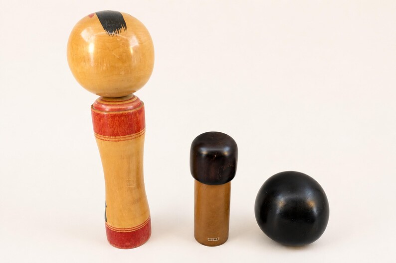 K&ouml;nnte beinhalten: Drei Holzobjekte, darunter eine gro&szlig;e Kokeshi-Puppe mit rundem Kopf und rot bemaltem K&ouml;rper, ein kleines zylindrisches Objekt mit dunkelbrauner Kappe und eine glatte schwarze Kugel. Die Gegenst&auml;nde sind in einer Reihe vor einem schlichten Hintergrund angeordnet.