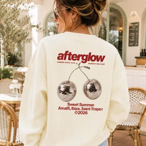 Peut inclure: Un sweat-shirt à col rond couleur crème avec un motif au dos portant le texte "afterglow", "SUMMER SOCIAL CLUB", "MIDNIGHT DISCO EDIT", "Sweet Summer", "Amalfi, Ibiza, Saint-Tropez" et "©2026" au-dessus et au-dessous de deux cerises boules à facettes.