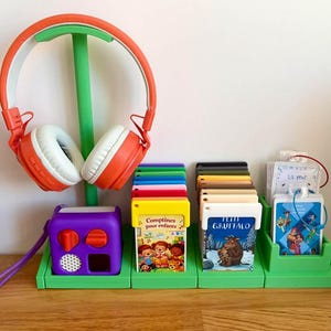 Puede incluir: Un soporte de almacenamiento de plástico verde con un gancho para auriculares naranjas y blancos, un reproductor de audio morado con correa, y varias ranuras que contienen tarjetas multimedia rectangulares y pequeños mazos de cartas.
