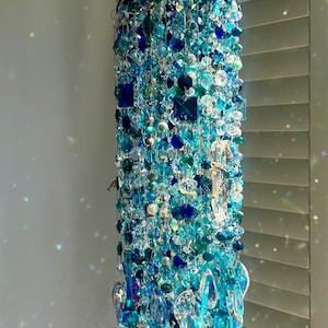 Pode incluir: Um sino de vento decorativo suspenso, com uma cascata de cristais e contas azuis e transparentes. A parte superior apresenta um design floral turquesa, com o corpo do sino de vento composto por cristais azuis e transparentes de várias formas e tamanhos.