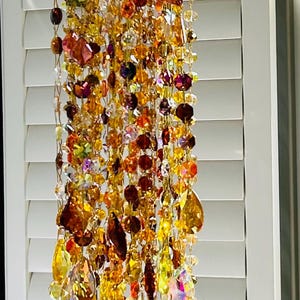Pode incluir: Um ornamento decorativo de cristal suspenso com um anel cor de cobre no topo. O ornamento apresenta fios de cristais âmbar, dourados e transparentes, com várias formas e tamanhos. Os cristais são suspensos em frente a uma persiana branca.