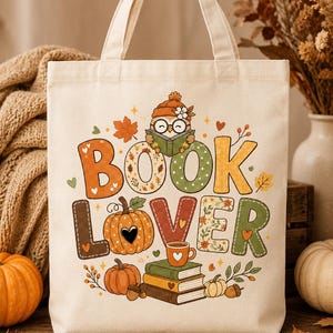 May include: A cream-colored canvas tote bag featuring a colorful graphic print with the text "BOOK LOVER". The design includes an owl wearing a hat and glasses reading a book, pumpkins, stacked books, a coffee mug, and autumn leaves in orange, green, and brown.