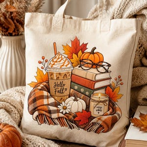 May include: A cream-colored canvas tote bag featuring a colorful graphic of an iced coffee with the text Hello Fall, a stack of books, a pumpkin, a lit candle labeled cozy vibes, and a plaid blanket surrounded by orange and yellow autumn leaves.