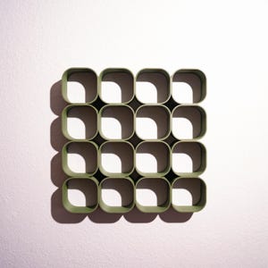 May include: A square wall-mounted storage unit featuring a grid of sixteen open compartments with rounded corners. The structure is finished in a matte olive green color, creating a repeating pattern of hollow squares that cast deep shadows.