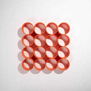 May include: A bright orange storage organizer featuring a grid of sixteen circular compartments arranged in a four-by-four pattern. The structure is made of connected cylindrical tubes, creating a uniform honeycomb-like appearance for holding items.