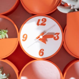 Könnte beinhalten: Eine Wanduhr mit weißem, kreisförmigem Zifferblatt und fetten orangefarbenen Ziffern 3, 6, 9 und 12. Die Uhr hat orangefarbene Zeiger und ist in ein modulares, orangefarbenes, kreisförmiges Rastersystem mit kleinen Pflanzgefäßen integriert.