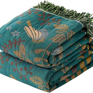 May include: A folded teal throw blanket featuring a woven pattern of birds, leaves, and floral motifs in shades of gold, brown, and grey. The blanket has a textured surface and is finished with a decorative green fringe along the top edge.