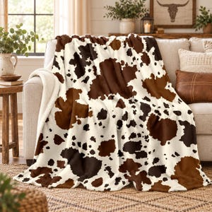 May include: A plush throw blanket featuring a brown and cream cow print pattern. The blanket has a soft, textured surface with irregular dark brown and light brown patches scattered across a cream-colored background, draped over a sofa.