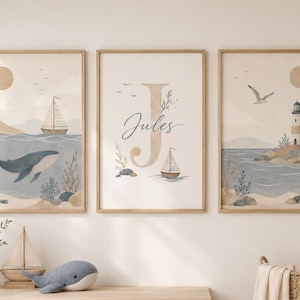 Könnte beinhalten: Drei gerahmte Wandbilder mit nautischem Motiv, bestehend aus einem Wal, einem Segelboot und einem Leuchtturm. Das mittlere Bild zeigt einen großen Buchstaben J und den Namen Jules in Schreibschrift. Die Farbpalette umfasst Beige, Blau und Weiß.