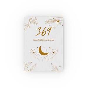 May include: A white journal with gold-coloured line art featuring hands, leaves, stars, and a crescent moon. The text 369 Manifestation Journal is printed in gold in the centre. The cover design includes delicate botanical illustrations and celestial symbols.