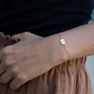 May include: A delicate gold chain bracelet with a small white skull charm. The bracelet is worn on a person's wrist.