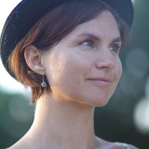 May include: A woman with short brown hair wearing a black hat and a silver earring with a blue stone.