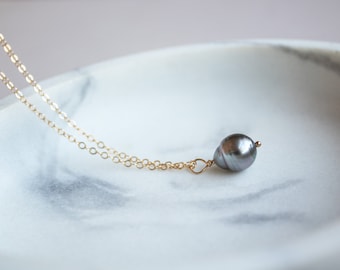 May include: A delicate gold chain necklace with a single, large, dark gray pearl pendant.