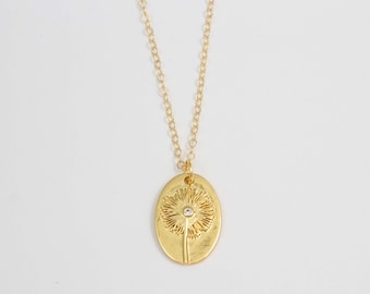 Puede incluir: Un collar de oro con una cadena delicada y un colgante ovalado que presenta un diseño de diente de león detallado con una pequeña piedra transparente en el centro.