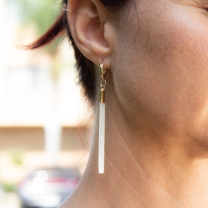May include: A pair of gold hoop earrings with a long, white, rectangular pendant hanging from each hoop.