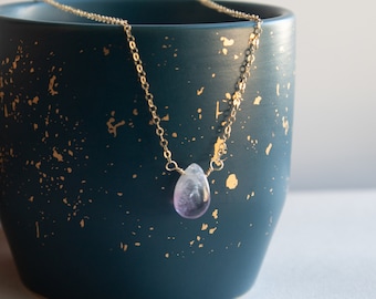 Puede incluir: Un delicado collar de cadena dorada con un colgante de amatista en forma de lágrima. El colgante es de color púrpura claro y está suspendido de una fina cadena dorada. El collar se muestra sobre un recipiente de cerámica azul oscuro con motas doradas.