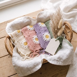 May include: A collection of four crocheted bonnets in cream, dusty pink, lavender, and sage green, each featuring a central white daisy motif with a yellow center. The bonnets have scalloped edges and thin ties, accompanied by a small white gift tag.