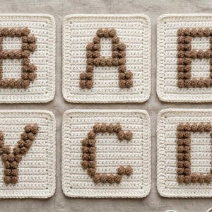 May include: Six square crochet patches arranged in two rows, each featuring a cream-colored base with a brown bobble-stitch letter. The letters spell out B, A, B, Y, C, D. A wooden crochet hook is visible in the bottom right corner of the frame.