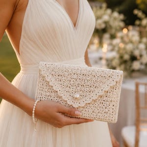 May include: A cream-coloured envelope-style clutch bag featuring an intricate crochet pattern with a scalloped edge flap. The clutch is secured with a single, centred, round white pearl button, providing a textured and detailed appearance.