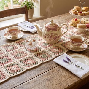 May include: A rectangular crochet table runner featuring a repeating granny square pattern in cream, sage green, and dusty rose. The runner is laid out on a wooden table with a floral porcelain teapot, teacups, a tiered dessert stand, and napkins.