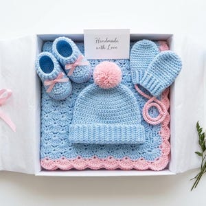 Könnte beinhalten: Ein Baby-Geschenkset in einer weißen Schachtel mit rosa Schleife, bestehend aus einer hellblauen Häkeldecke mit rosa Bogenkante, einer passenden hellblauen Mütze mit rosa Bommel, Babyschuhen mit rosa Schleifen und Fäustlingen. Karte: Handmade with Love.