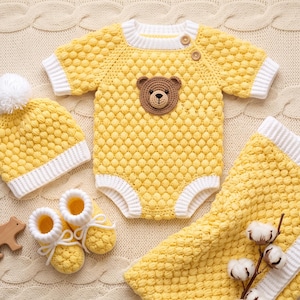 May include: A yellow and white knitted baby set featuring a short-sleeved romper with a brown bear face appliqué, a matching beanie with a white pom-pom, a pair of booties with white laces, and a blanket, all with a textured bobble stitch pattern.