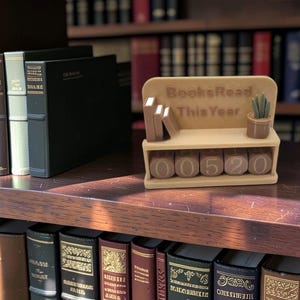 Peut inclure: Une étagère de suivi de lecture en plastique beige avec le texte BooksRead This Year, comprenant trois petits blocs en forme de livres, une plante miniature en pot et quatre blocs numériques rotatifs affichant 00520, sur une étagère en bois.