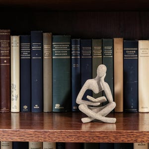 Pode incluir: Uma estatueta humana abstrata de cor bege, com textura granulada, sentada de pernas cruzadas e segurando um livro aberto. A figura tem uma cabeça lisa e sem traços, apoiada numa mão, com uma forma simples e um acabamento semelhante a pedra.