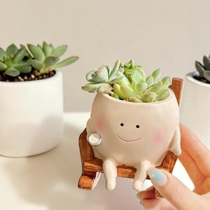 May include: A cream-colored ceramic planter shaped like a round character with a smiling face and pink cheeks, sitting in a small brown wooden rocking chair. The planter holds a green succulent plant, with two other plain white planters nearby.