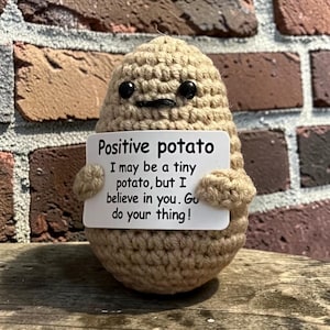 May include: A tan crocheted potato figure with two black bead eyes and a small stitched mouth. The figure holds a white rectangular sign that reads: Positive potato. I may be a tiny potato, but I believe in you. Go do your thing!