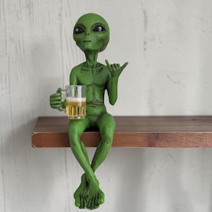 May include: A green alien figurine with large black eyes, sitting with legs crossed while holding a clear glass mug filled with a golden liquid topped with white foam. The alien has one hand raised in a shaka gesture, featuring a smooth texture.