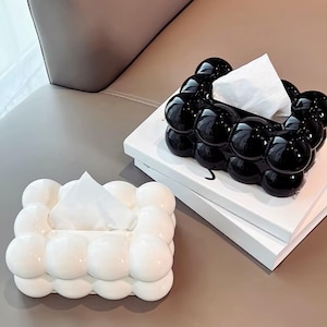 Può includere: Due scatole rettangolari per fazzoletti, una in nero lucido e una in bianco lucido, caratterizzate da una texture tridimensionale a bolle o sfere sulle superfici esterne. Ogni scatola presenta un'apertura rettangolare centrale.