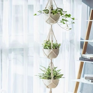 May include: A three-tier hanging planter featuring off-white macrame cord netting holding three smooth, round, off-white ceramic pots. Each pot contains a green houseplant with varying leaf shapes and sizes, suspended vertically in a column.