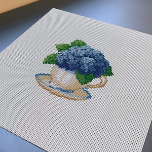 May include: A cross-stitch embroidery piece on white Aida fabric featuring a teacup filled with a cluster of blue hydrangea flowers and green leaves. The teacup is light tan with a blue rim and handle, centred on the square fabric canvas.