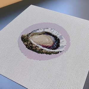 May include: A square needlepoint canvas featuring a centered, circular design of an open oyster shell. The shell is rendered in shades of brown, cream, and dark purple, set against a soft, pale lavender circular background on a white grid.